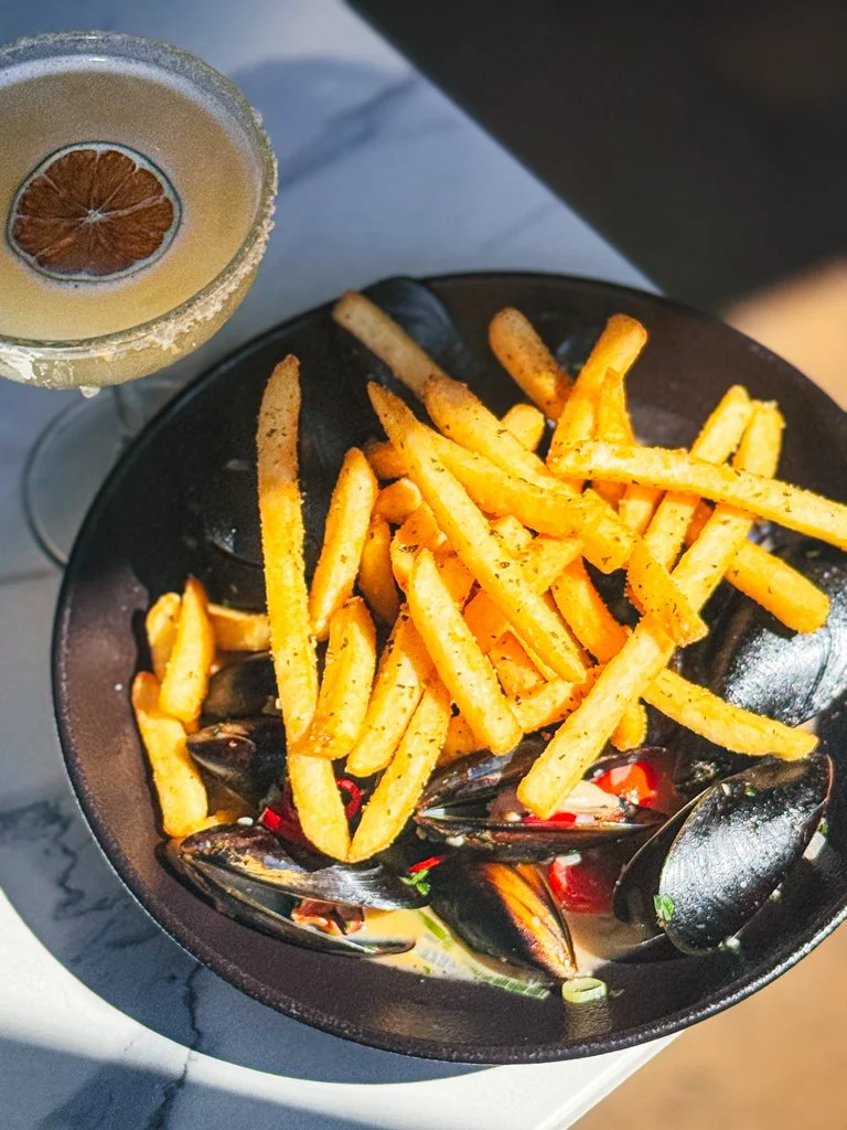 French fries with mussels in a black bowl, served with a beverage that has a lemon slice and salt rim.