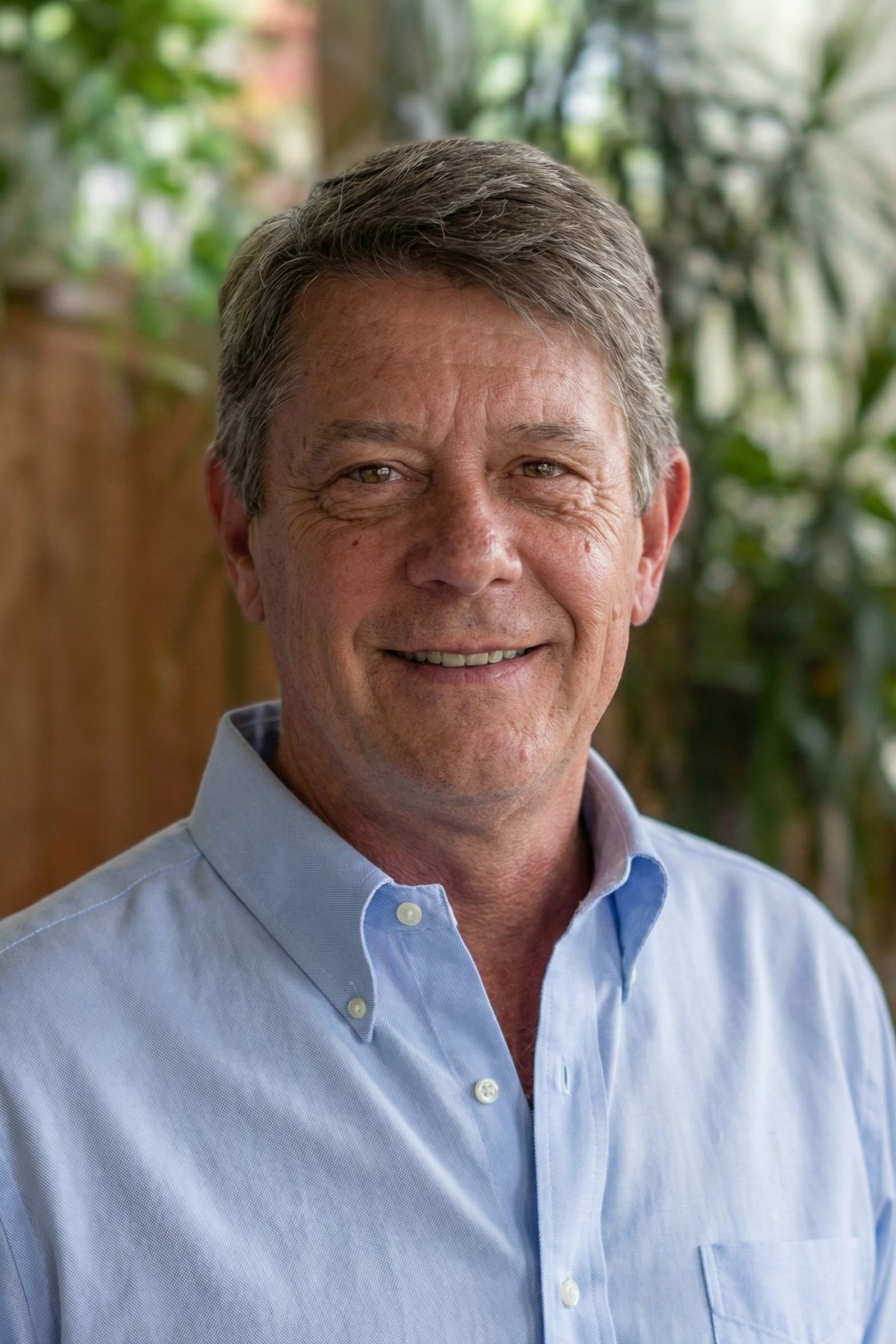 A smiling middle-aged man with short gray hair wearing a light blue button-up shirt, standing indoors with a background of green plants and natural light.