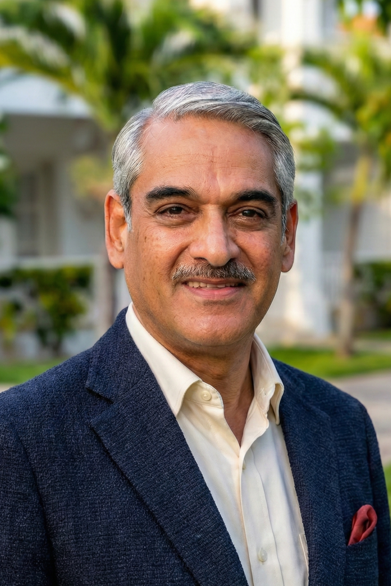 A middle-aged man with gray hair and a mustache, wearing a navy blazer and cream-colored shirt, smiling outdoors with trees and a house in the background.