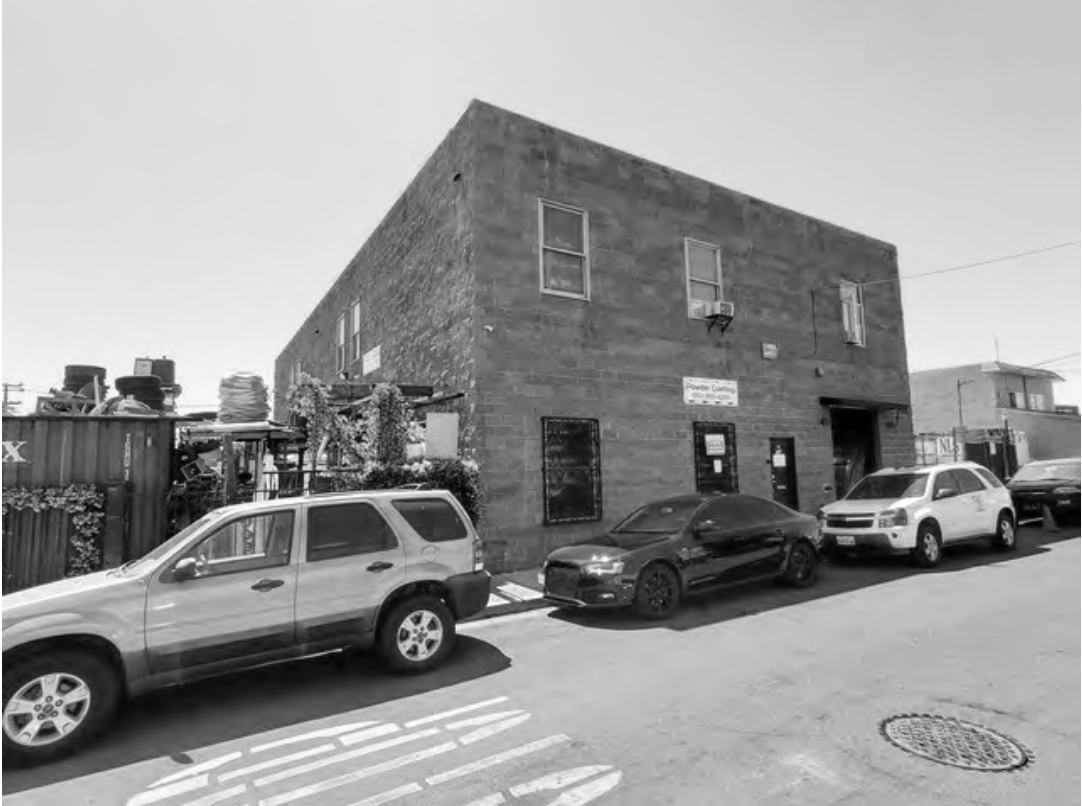 Black and white photo of a small brick commercial building with a sign, parked cars in front, and a fence with plants to the side.