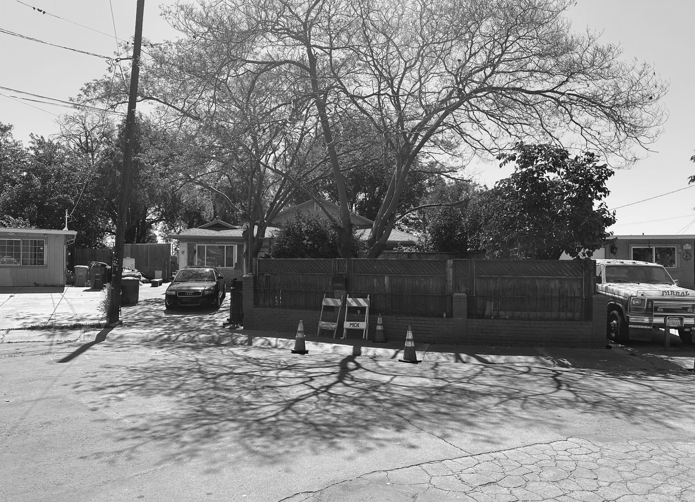 A residential street with a house behind a wooden fence, several parked cars, and trees casting shadows on the pavement.