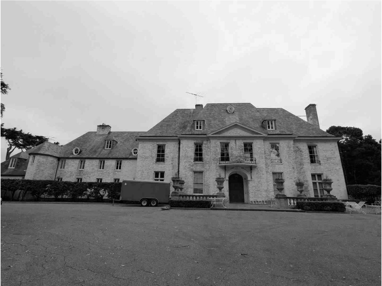 Black and white photo of a large, historic mansion with multiple windows and a central entrance, surrounded by a driveway and bushes.
