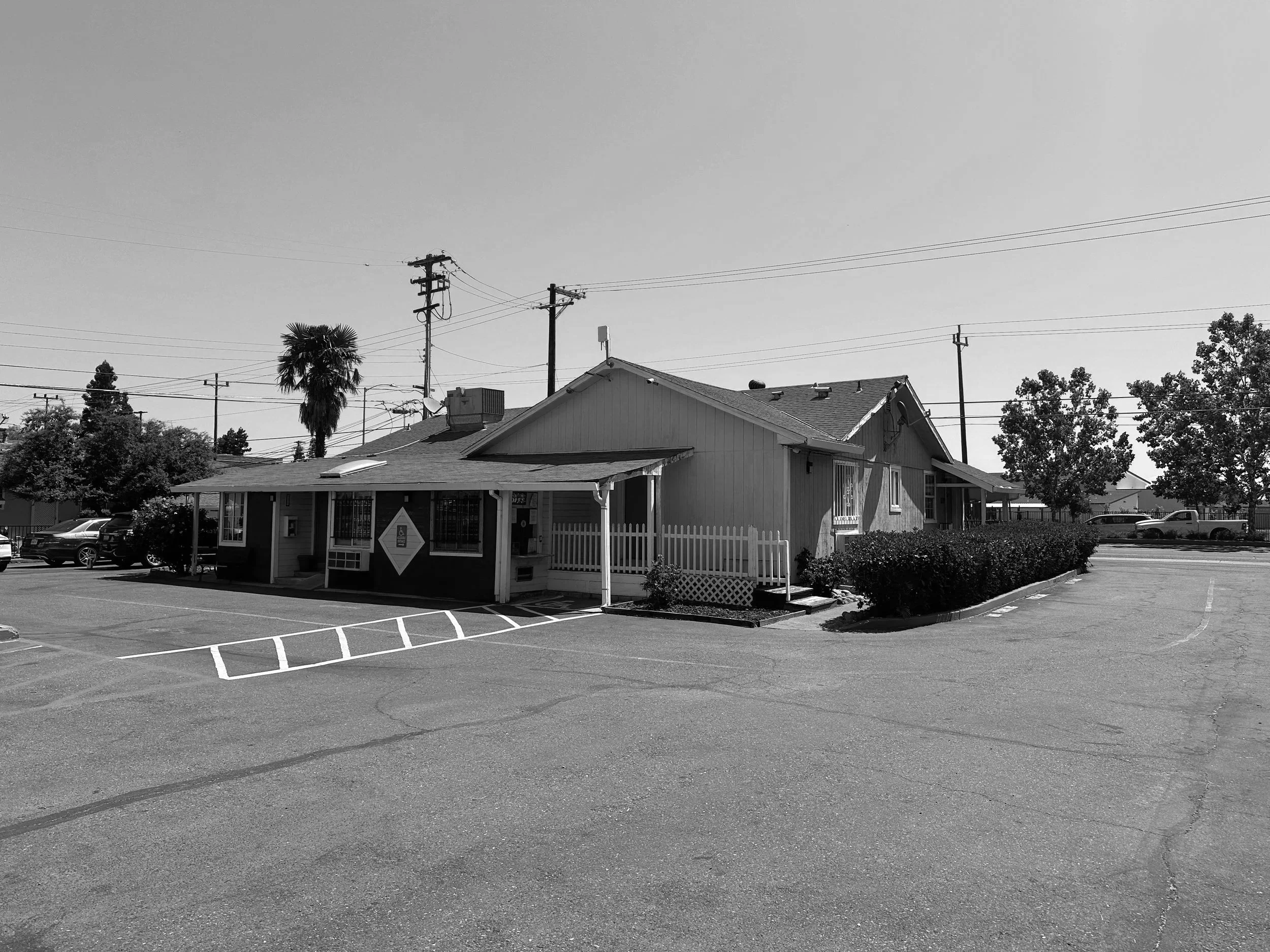 A black and white photo of a small building with a parking lot in front, surrounded by trees and utility poles.