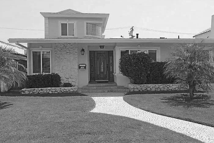 A two-story house with a front door, two windows, and a small upper window. The yard has a curved gravel path leading to the front steps, surrounded by trimmed bushes and palm trees.