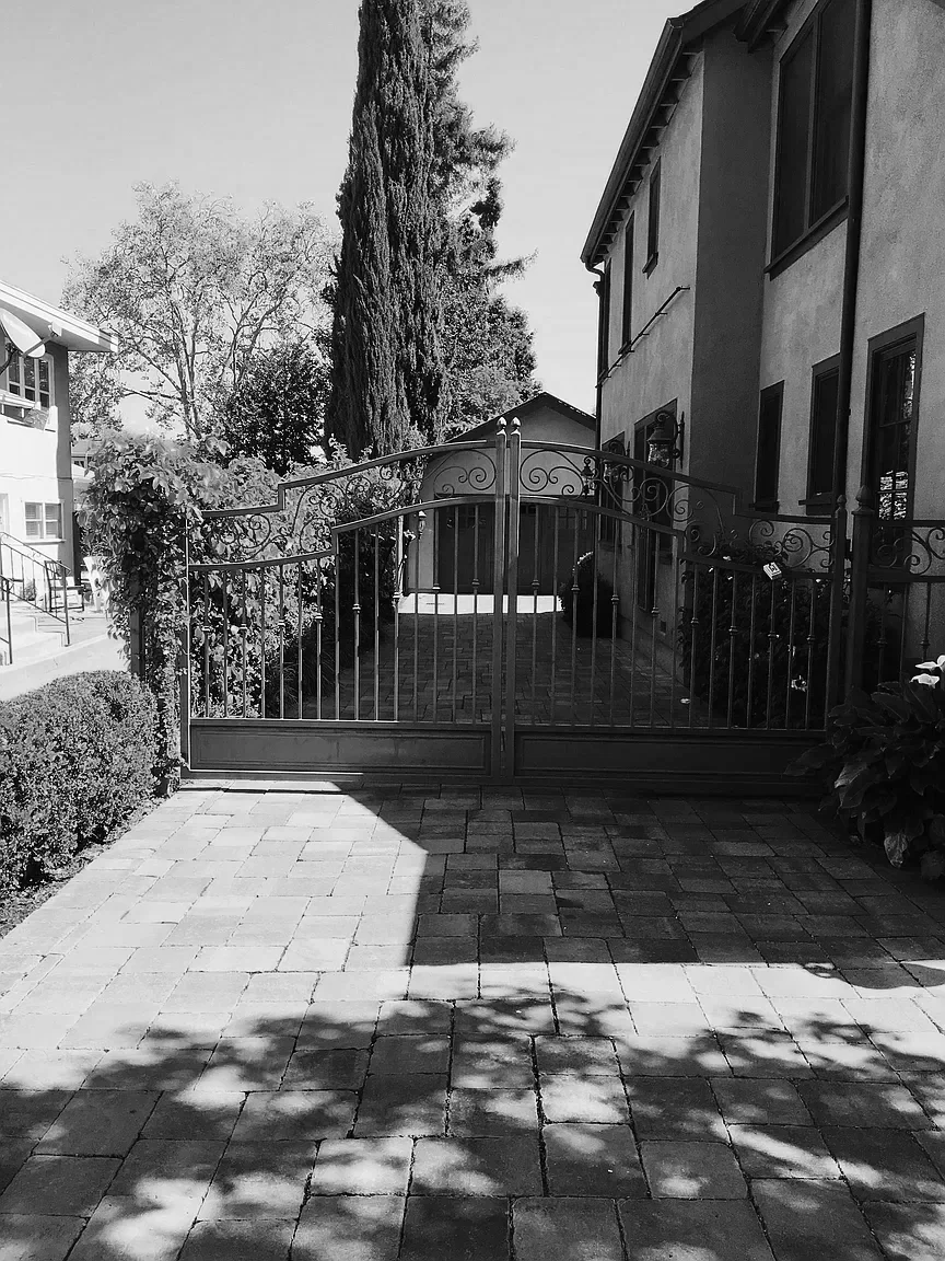 Residential backyard with a metal gate, brick driveway, and house on right side. Trees and bushes cast shadows on the driveway.