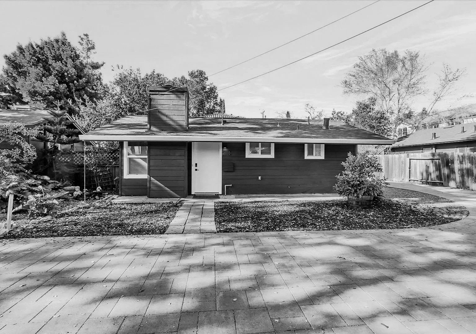Black and white photograph of a single-story house with a gabled roof, a central door, and three small windows. The house has a small front yard with a paved walkway, and a larger patio toward the bottom of the image. There are trees and a wooden fence surrounding the yard.
