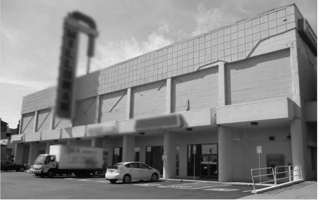 A two-story commercial building with a plain facade, parking spaces in front, and a UPS delivery truck and a white sedan parked outside.