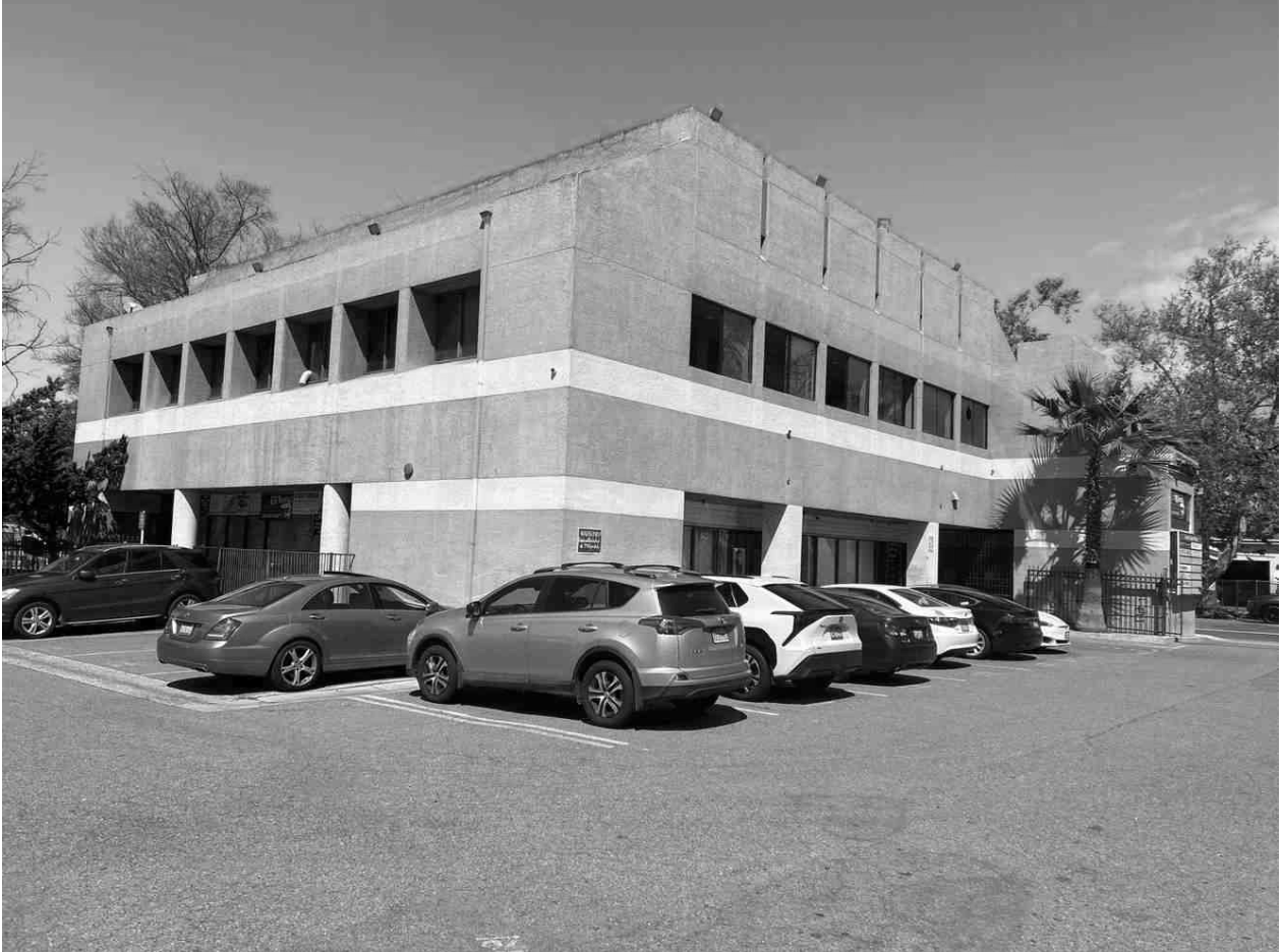 Black and white photo of a modern, multi-story parking lot with several parked cars in front, trees, and a fence.