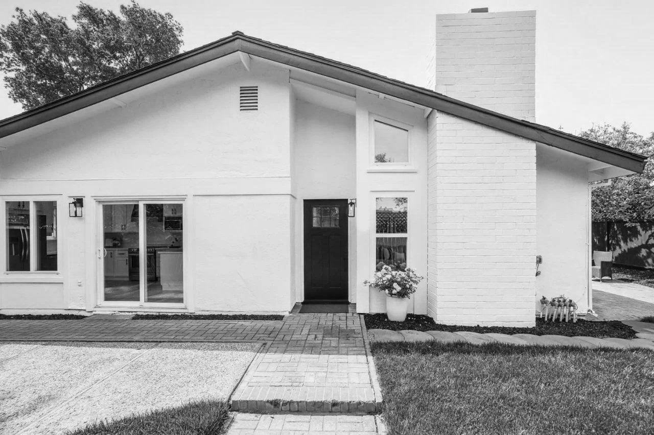 Black and white photo of a modern house with a sloped roof, brick chimney, large windows, and a front door with a flower pot nearby.