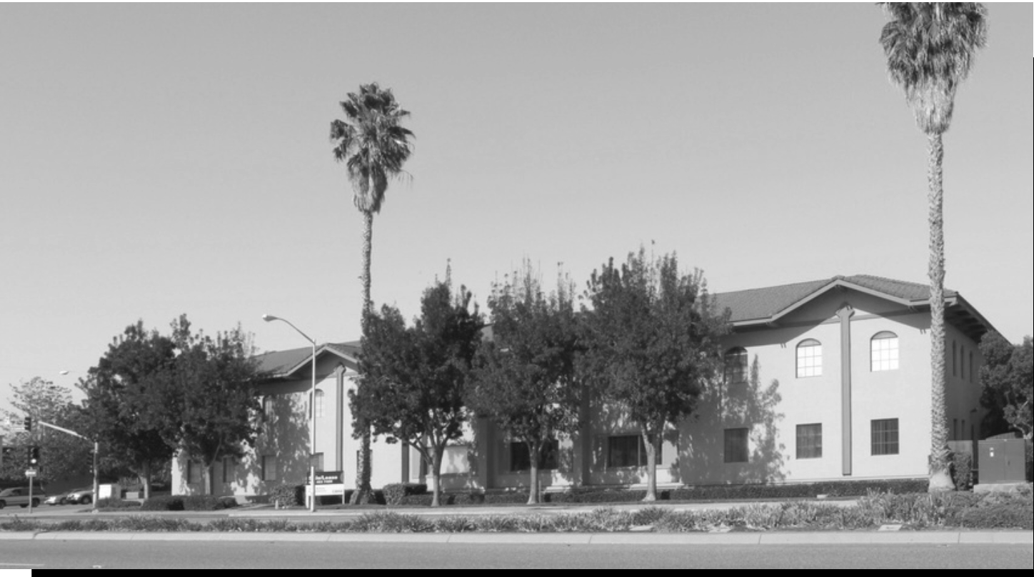 A residential building with multiple windows and a gabled roof, surrounded by tall palm trees and shorter trees, with streetlights and a road in the foreground.