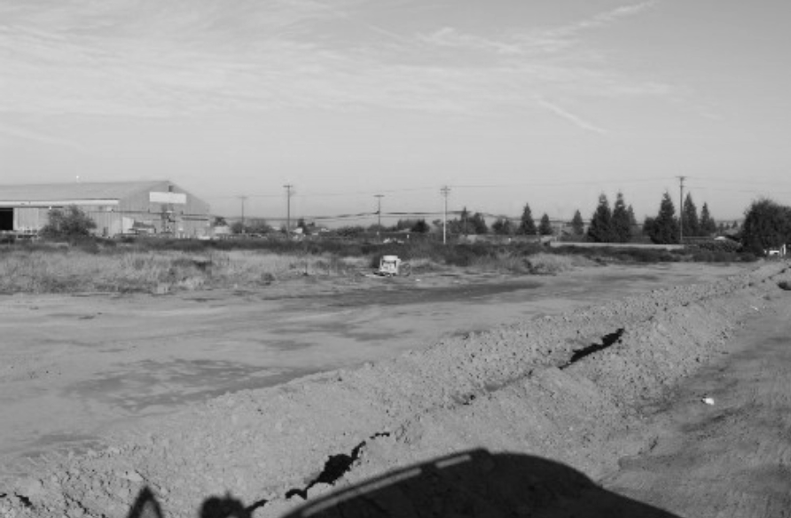 Black and white photo of an empty, uneven dirt road with cracks, and a small vehicle in the distance, with industrial and tree-lined buildings in the background under a sky with clouds.