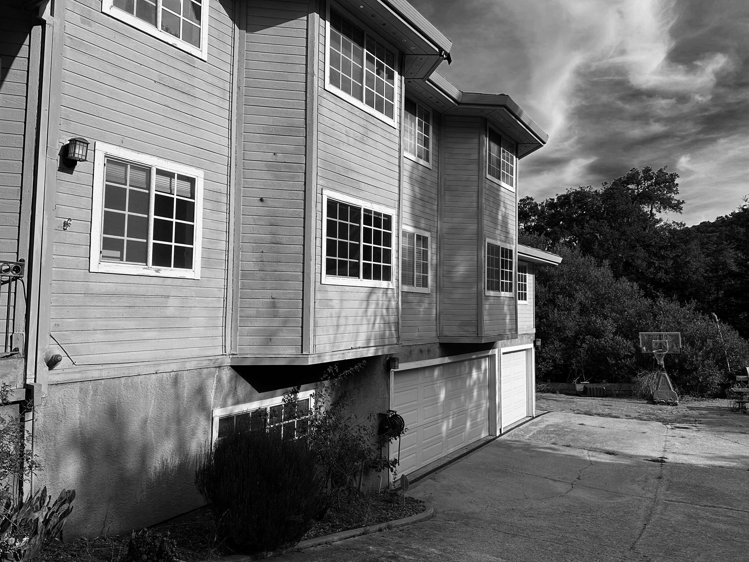 Black and white photo of a multi-story house with a garage door, several windows, and a basketball hoop in a yard with trees in the background.