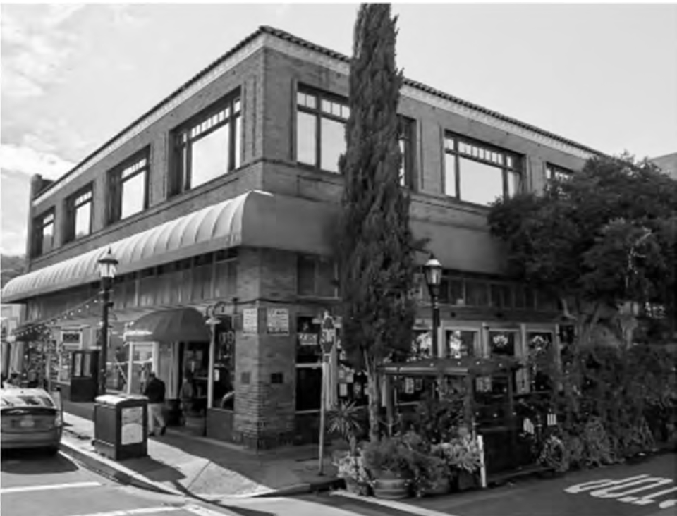 A black and white photo of a three-story building on a city street, with large windows, an awning over the sidewalk, and trees and street lamps in front.