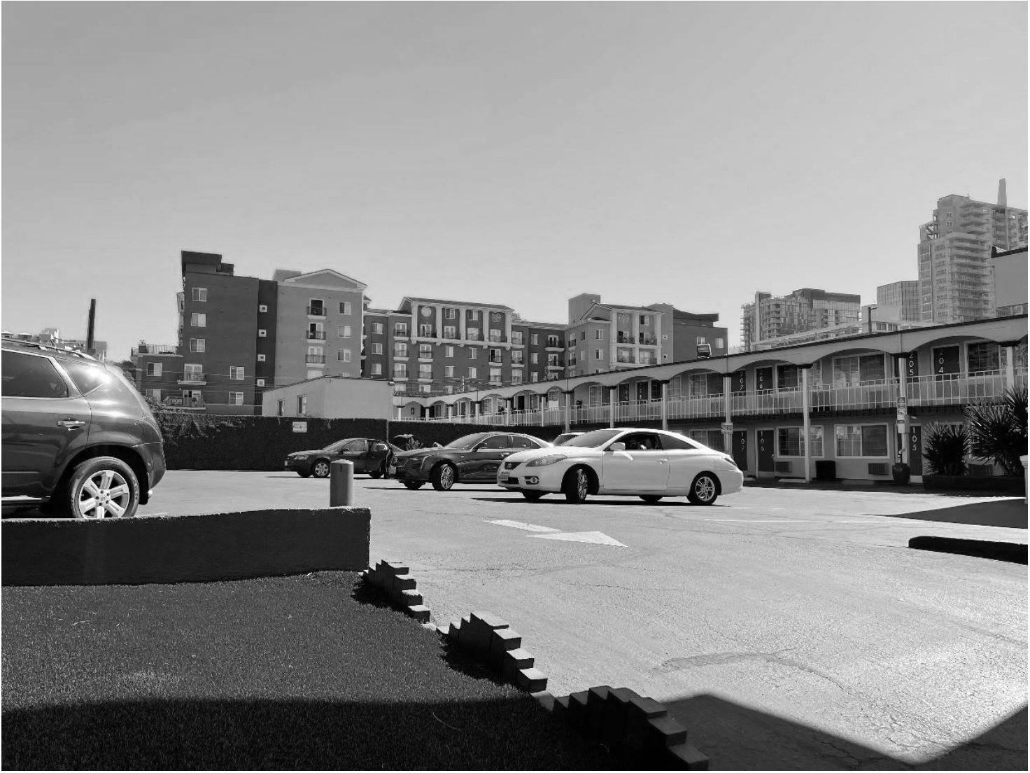 A parking lot with several parked cars, overview of mid-rise apartment buildings or condos in the background, in a cityscape with high-rise buildings further behind. Black and white.