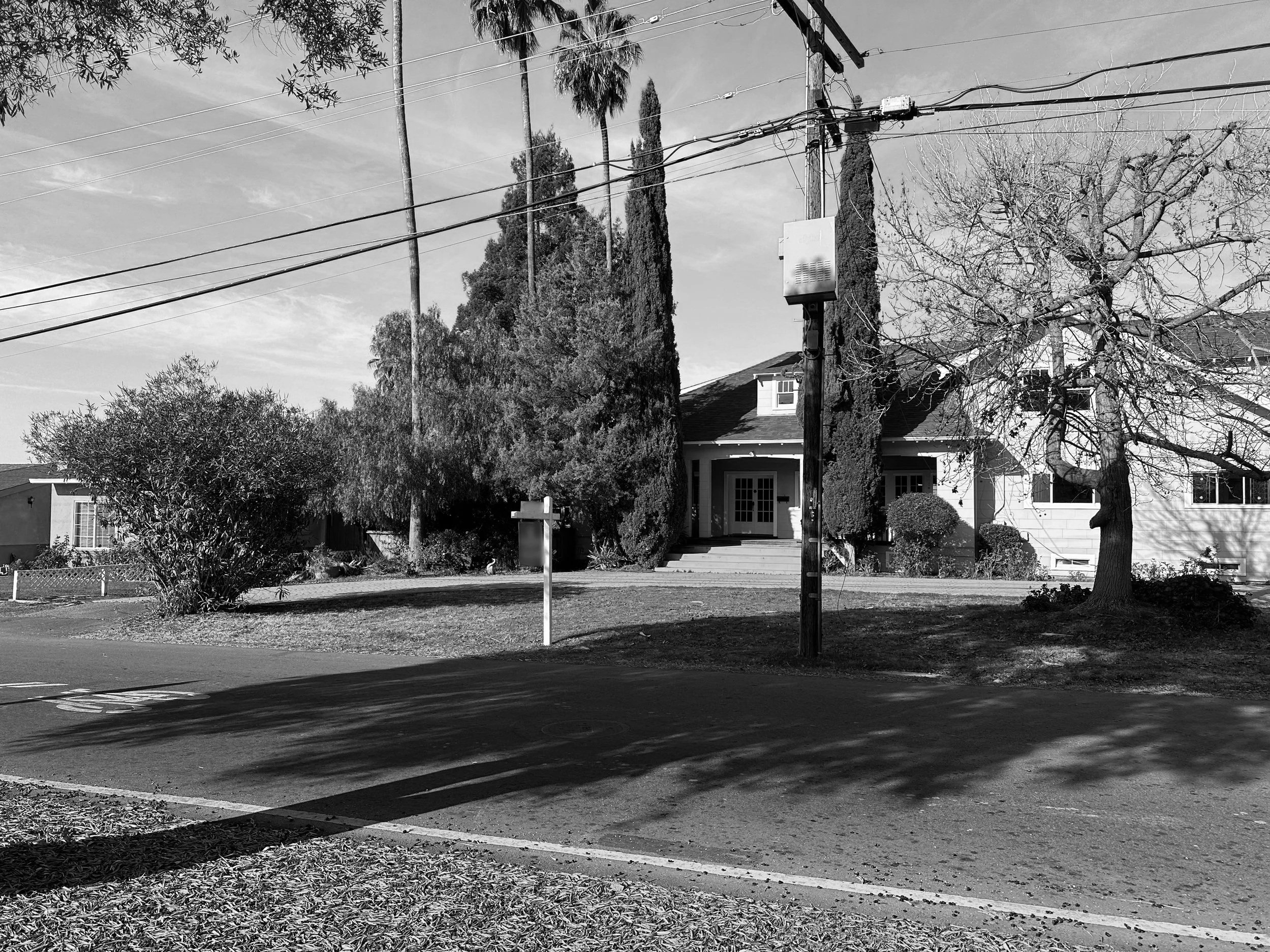 A black and white photo of a residential street corner featuring a house with a porch, trees, bushes, a mailbox, utility poles, and overhead power lines.