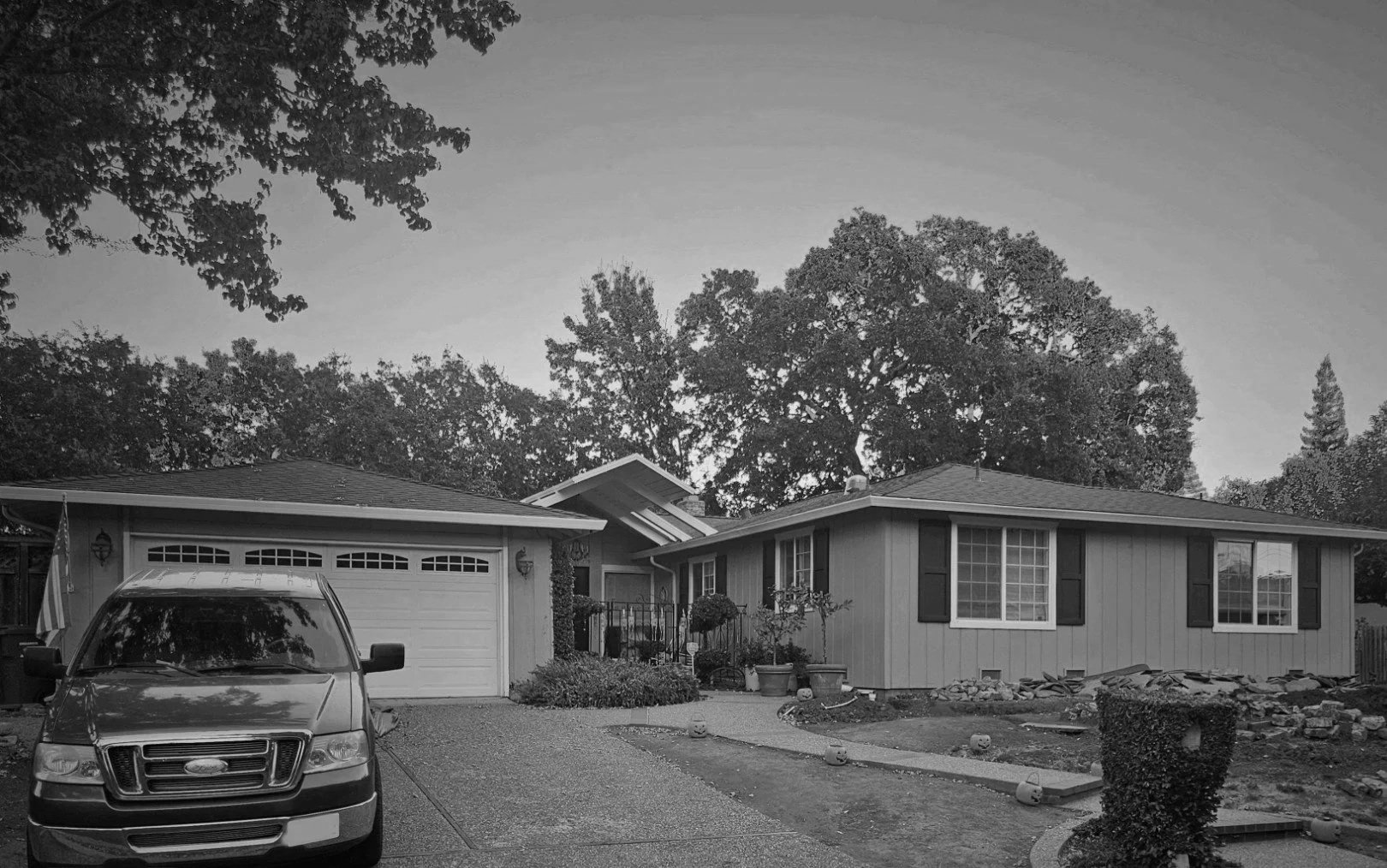 A suburban house with a driveway, a parked car, a garden with potted plants and small pumpkins, trees in the background, and a cloudy sky.