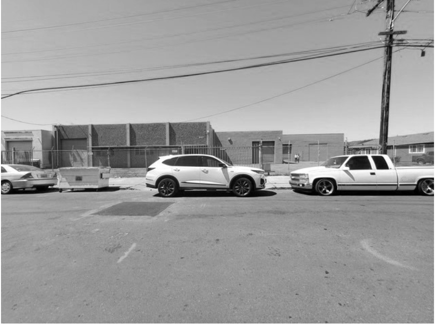 Street view with parked cars, utility pole, and buildings in the background, in black and white.