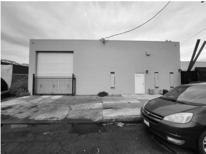 Gray building with a large roll-up garage door, a smaller standard door, and small windows, with cars parked in front on an urban street.
