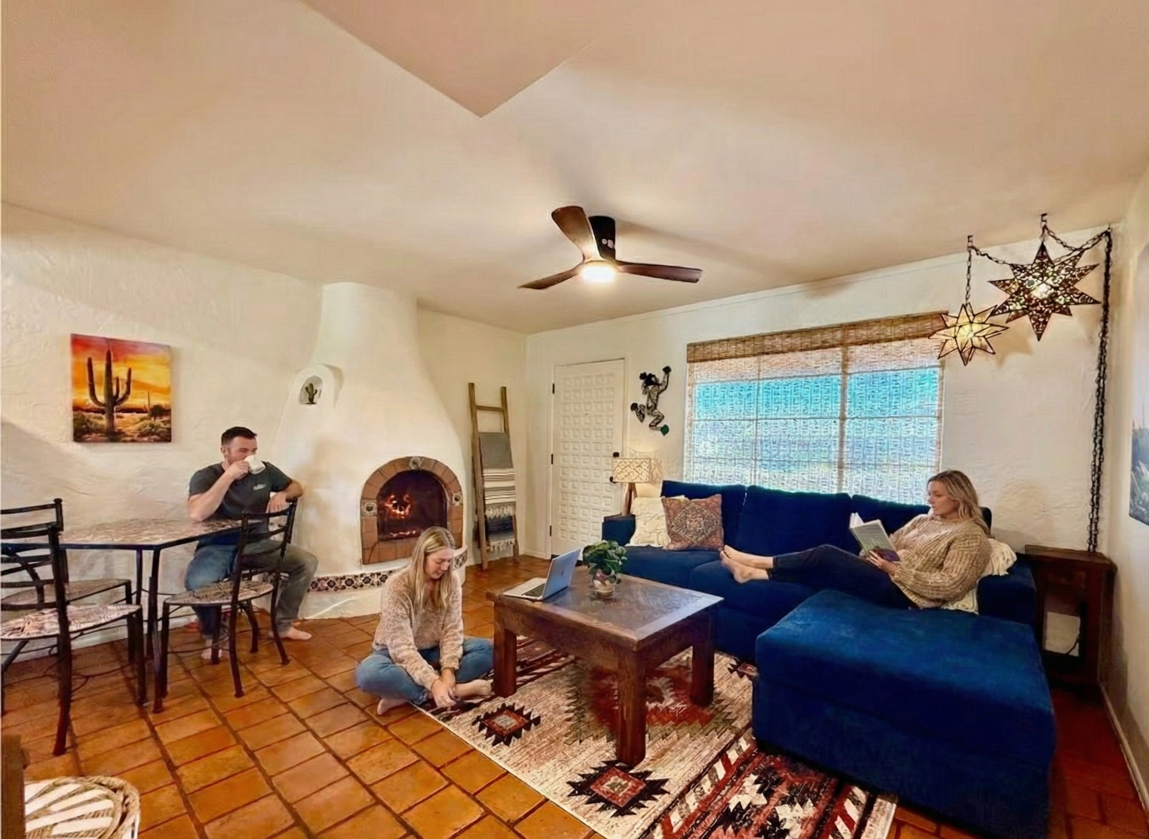 A cozy living room with three people. A woman is sitting on the floor, working on a laptop. Another woman is relaxing on a navy blue sofa, reading a book. A man is sitting at a dining table, drinking from a mug. The room features a fireplace, a ladder leaning against the wall, decorative star lanterns hanging from the ceiling, a patterned rug, and a window with bamboo blinds.