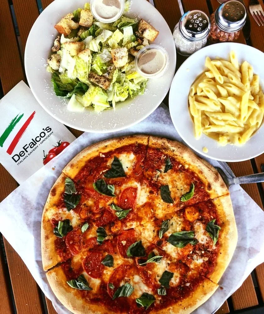 A meal consisting of a pepperoni pizza, a Caesar salad, and a side of fettuccine pasta. The pizza has cheese, tomato sauce, pepperoni, and basil leaves. The salad includes chopped romaine lettuce, croutons, and two small containers of dressing. There are salt and pepper shakers and a napkin with the DeFalco's Italian Eatery and Groceries logo on it.