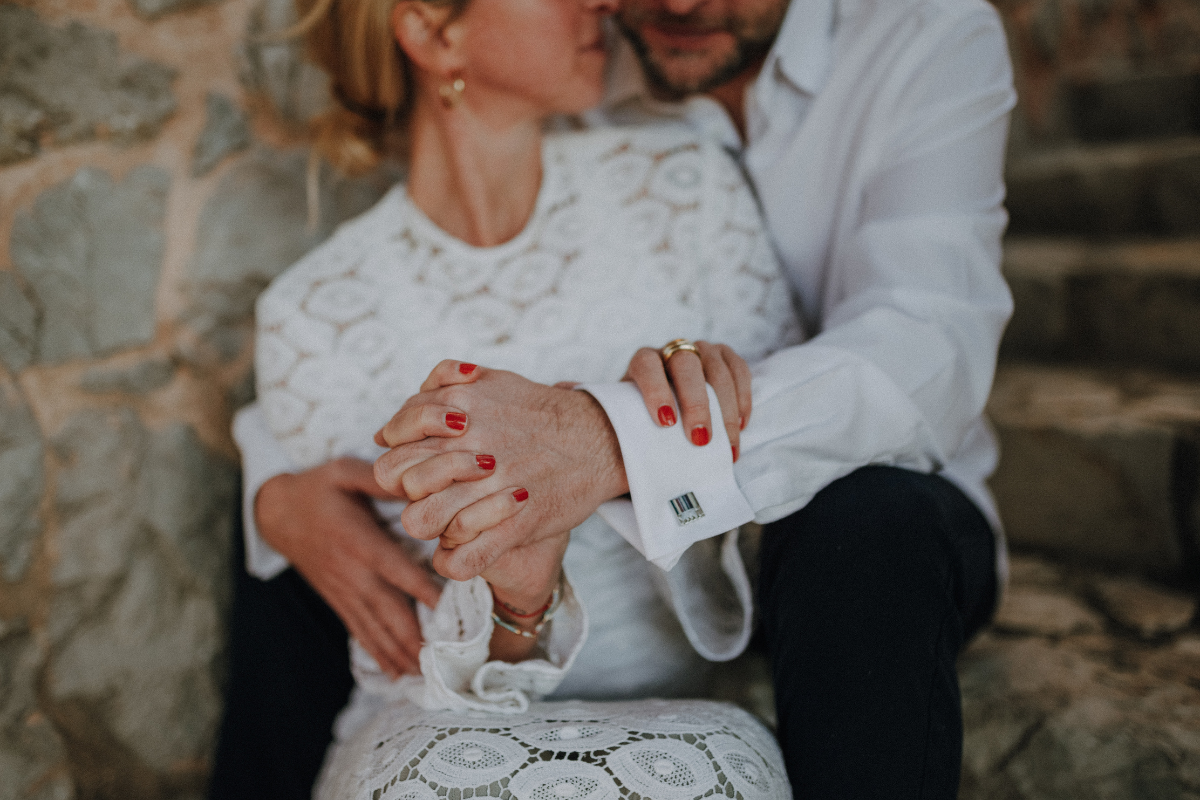 Ein älteres Paar umarmt sich liebevoll, beide tragen weiße Hemden, die Frau hat rote Nägel, sitzen auf einer Steintreppe vor einer Steinmauer.