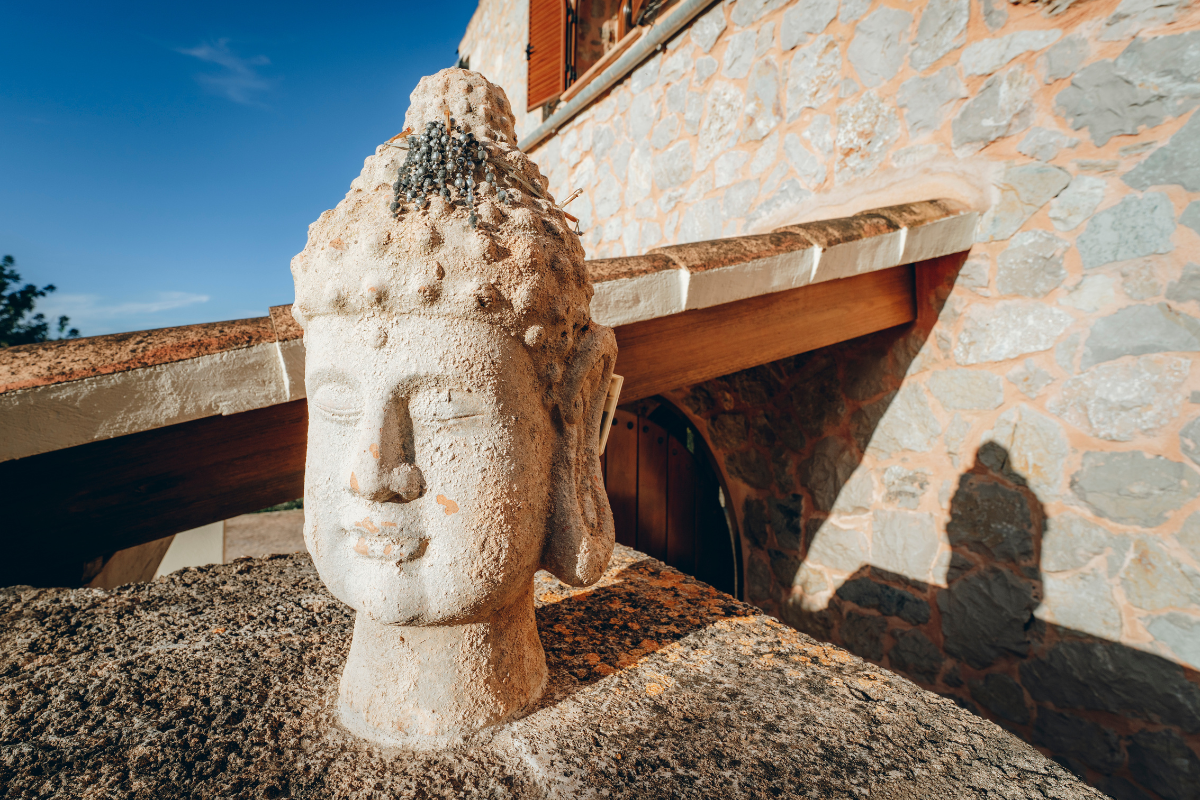 Steinerne Skulptur eines Buddha-Kopfes vor einem Steinmauer und einem Holzblock, mit der Sonne, die einen Schatten an die Wand wirft.