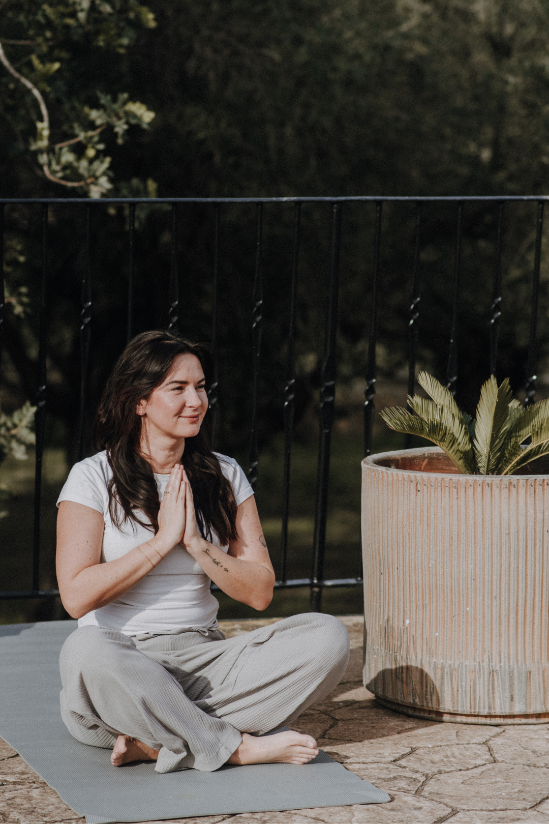 Eine Frau macht Yoga im Freien im Schneidersitz auf einer Yogamatte, neben einer großen Pflanzenterrinde.