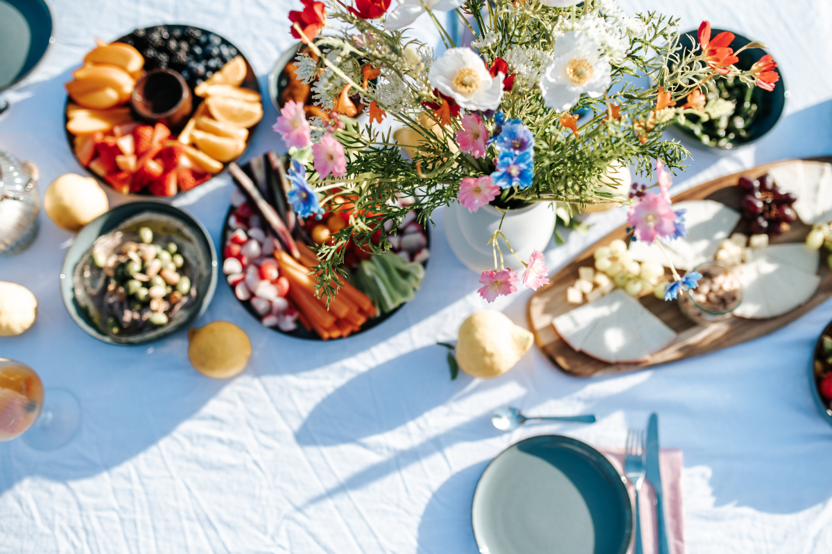 Bunt gedeckter Tisch mit einem großen Blumenstrauß, verschiedenen Käseplatten, Früchten, Naschereien und Snacks bei Tageslicht.