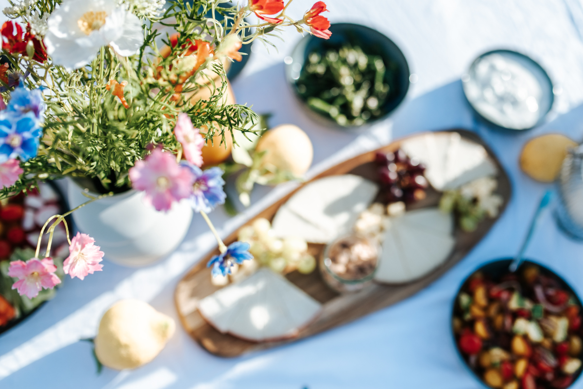 Blumenstrauß mit bunten Blumen und Tafel mit Käse, Obst und Joghurt auf weißem Tuch bei Sonnenlicht.
