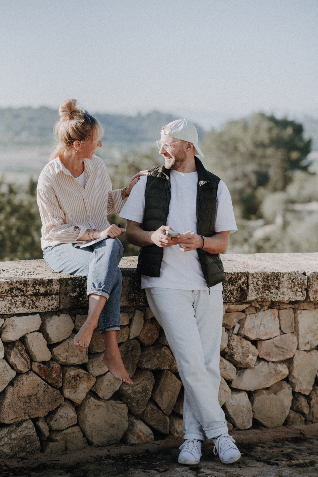 Ein Mann und eine Frau sitzen auf einer Steinmauer, lachen und unterhalten sich bei sonnigem Wetter im Freien, im Hintergrund Bäume und Hügel.