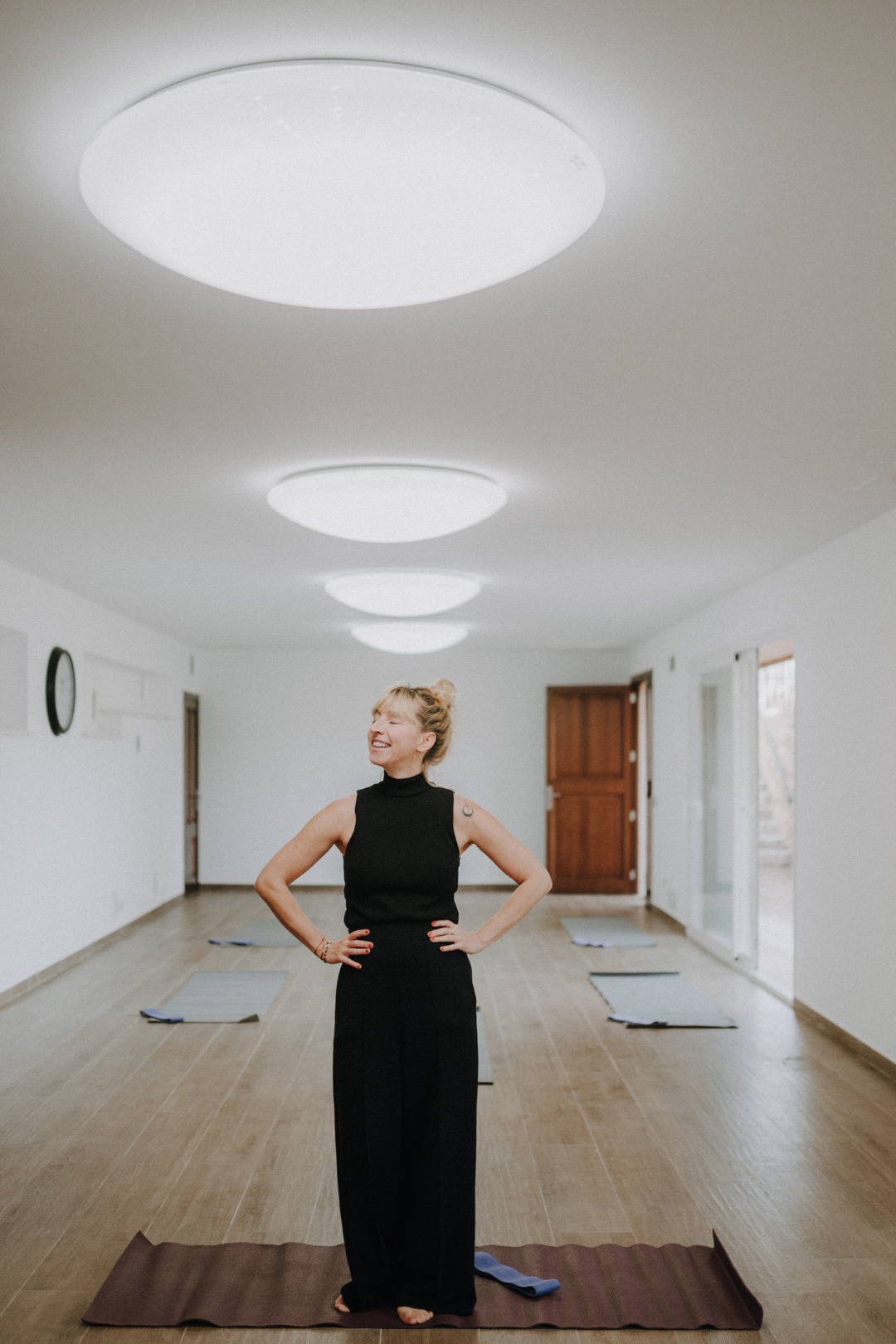 Eine lachende Frau steht auf einem Yogamatte in einem hellen Raum mit mehreren anderen Yogamatten. Der Raum hat weiße Wände, holzfarbene Dielen, und große Fenster. Über der Frau befinden sich runde, weiße Deckenleuchten.