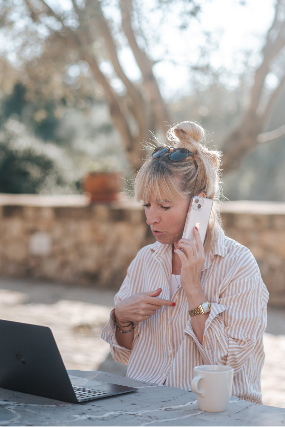 Eine blonde Frau mit Sonnenbrille auf dem Kopf telefoniert und auf ihr Smartphone zeigt während sie an einem Tisch sitzt, auf dem ein Laptop und eine Tasse stehen, im Freien bei sonnigem Wetter.
