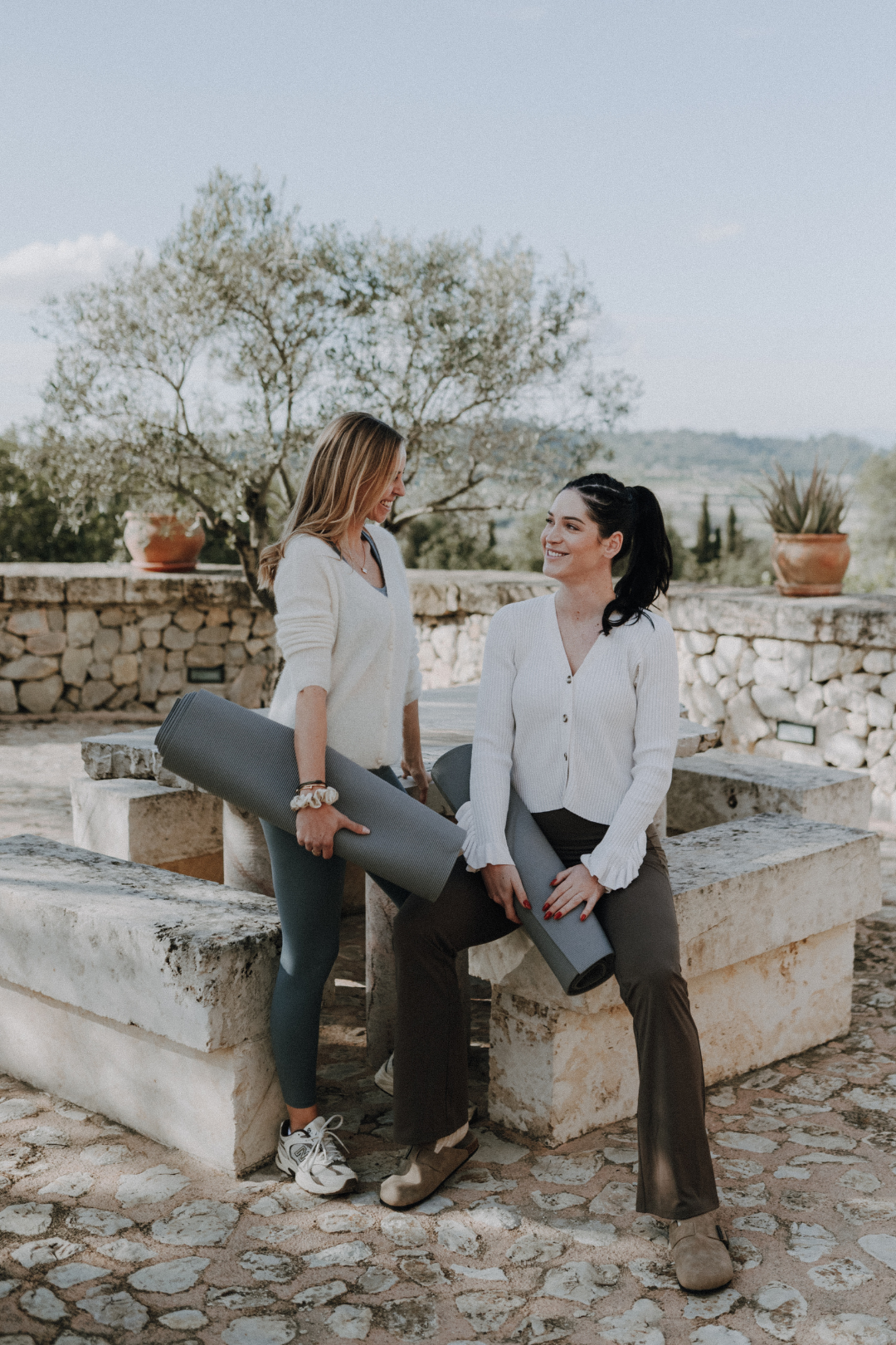 Zwei Frauen mit Yogamatten in einer outdoor Umgebung, lachend und unterhaltend.