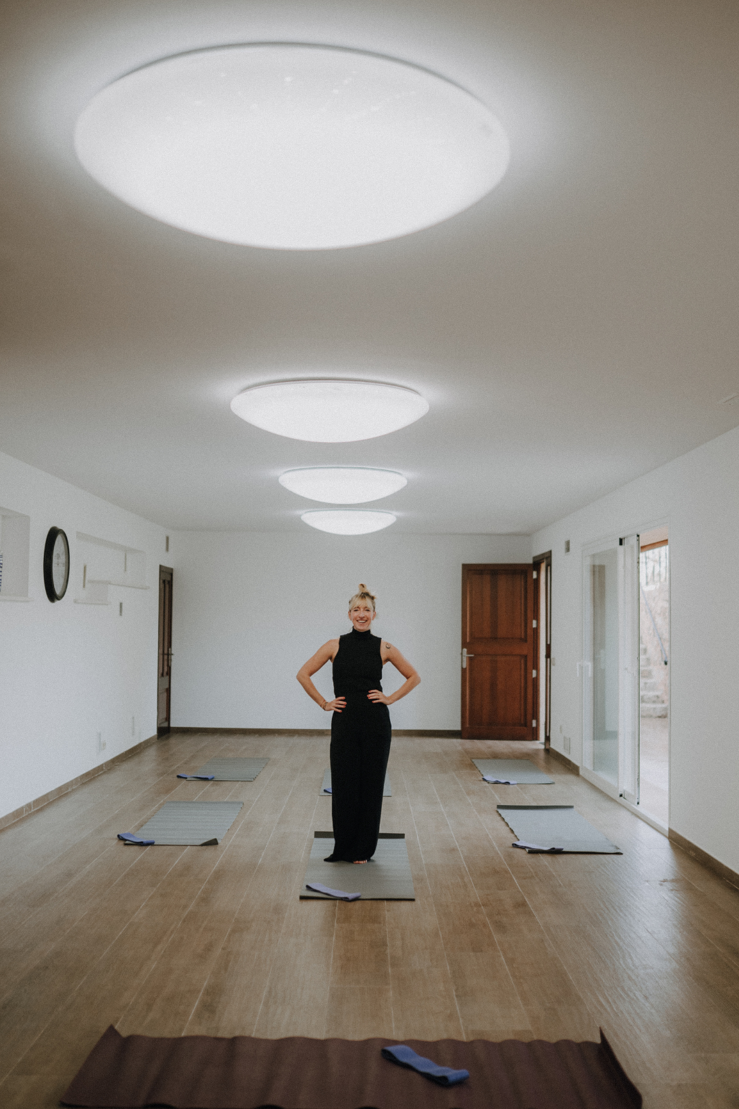 Eine Frau steht in einem leeren, hell erleuchteten Raum auf einer Yogamatte, umgeben von mehreren weiteren Matten, bereit für eine Yoga- oder Pilatesstunde.