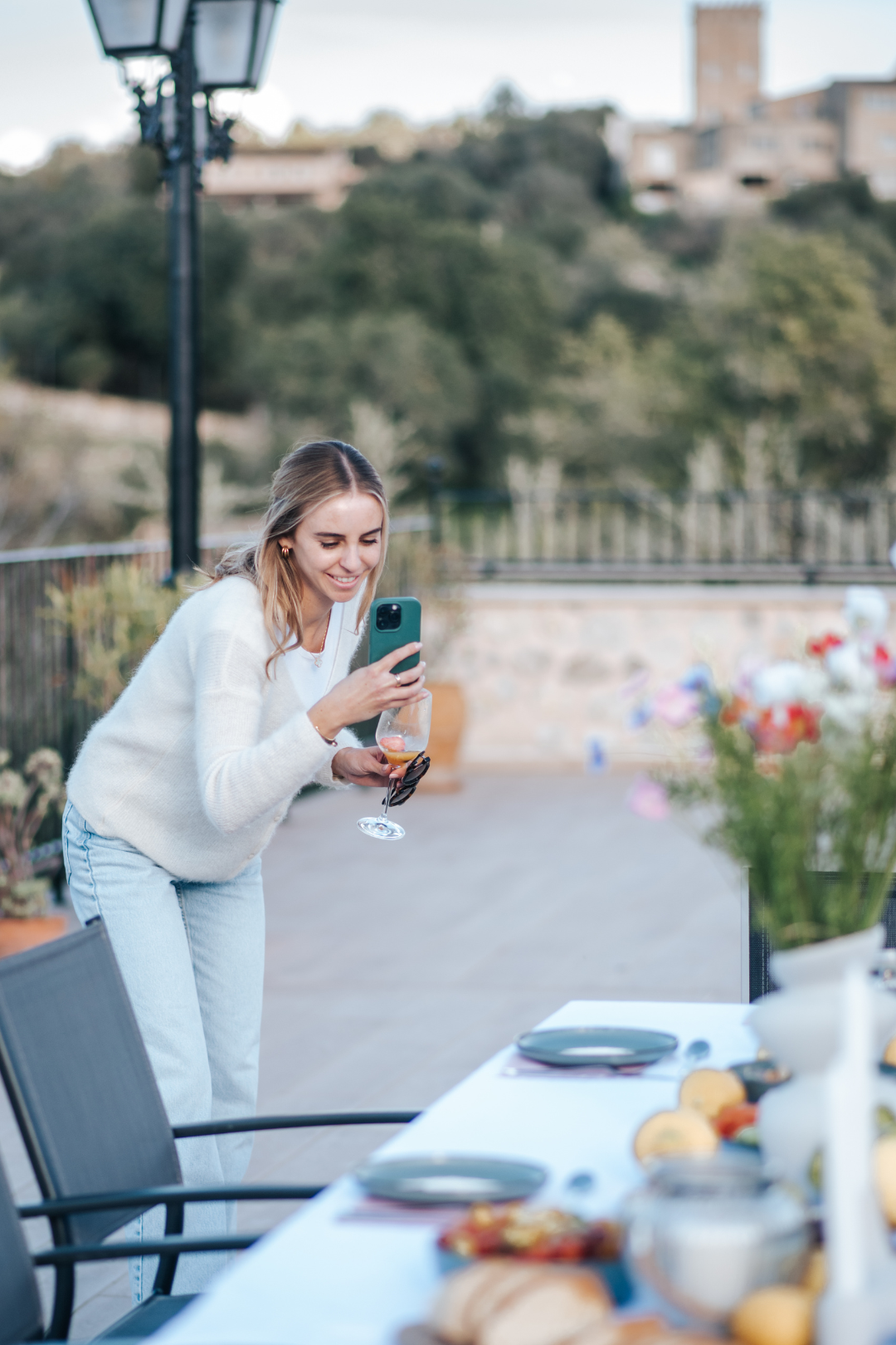 Junge Frau macht ein Selfie mit Smartphone bei einem Dinner im Freien bei Sonnenuntergang, mit einem Tisch voller Essen und Dekorationen im Vordergrund