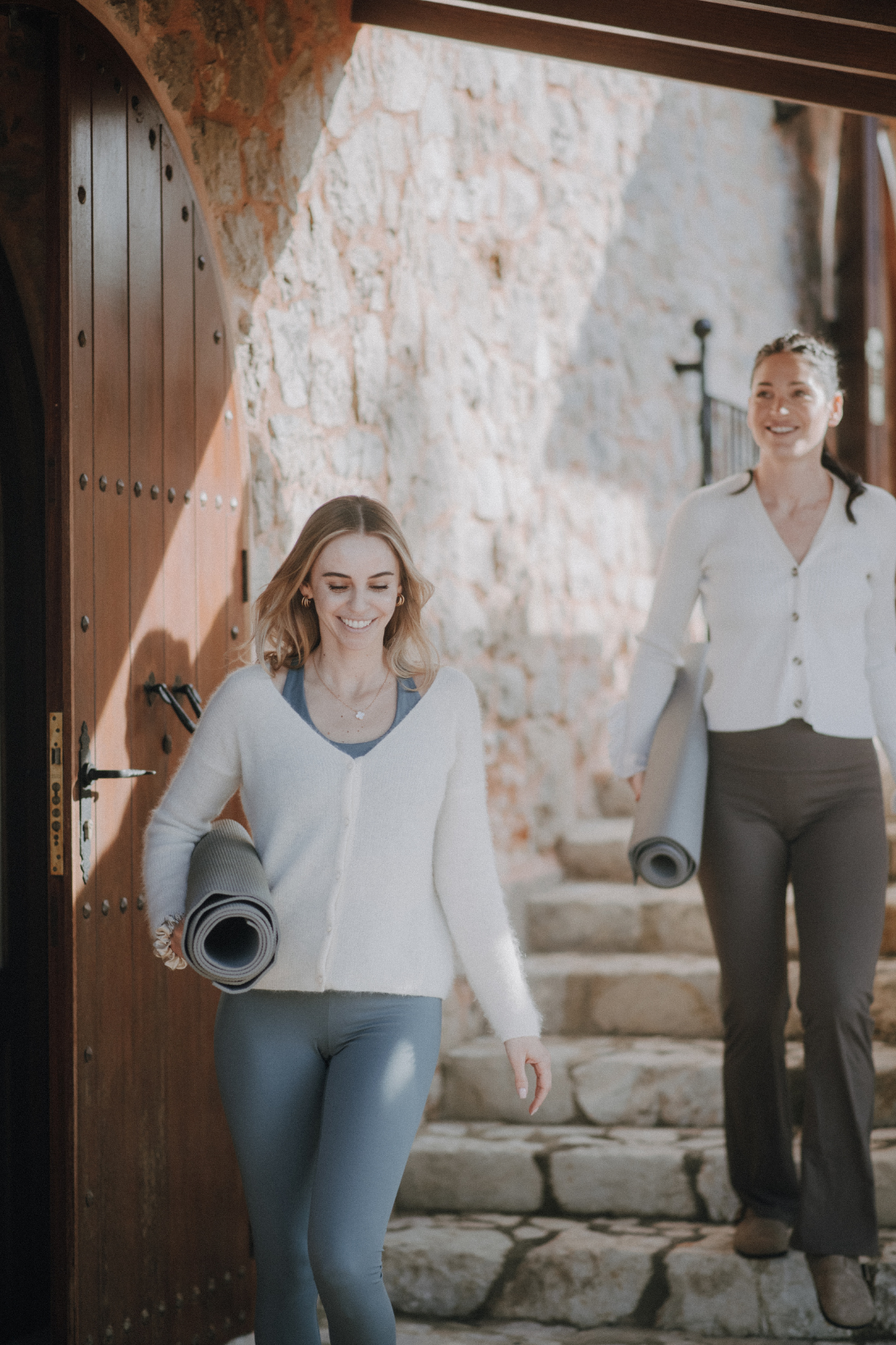 Zwei Frauen gehen die Steintreppe eines Hauses hinunter, tragen Yogamatten, lachen und haben Spaß.