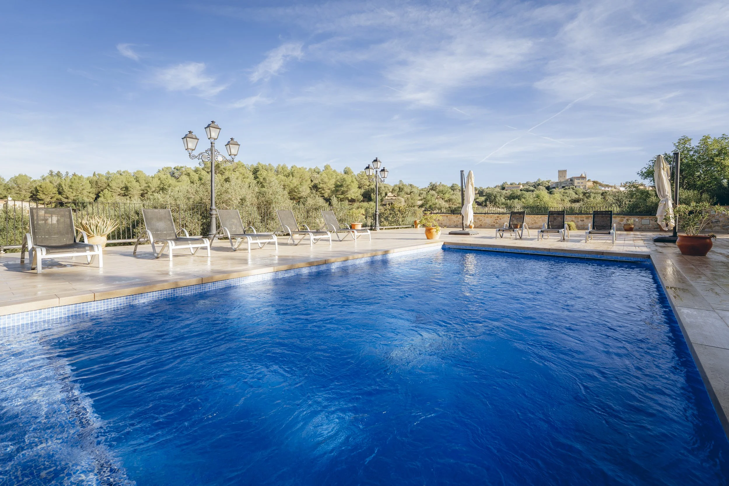 Blick auf einen Swimmingpool mit Poolliegen, Sonnenschirmen und Lampen, umgeben von Natur und einem blauen Himmel.