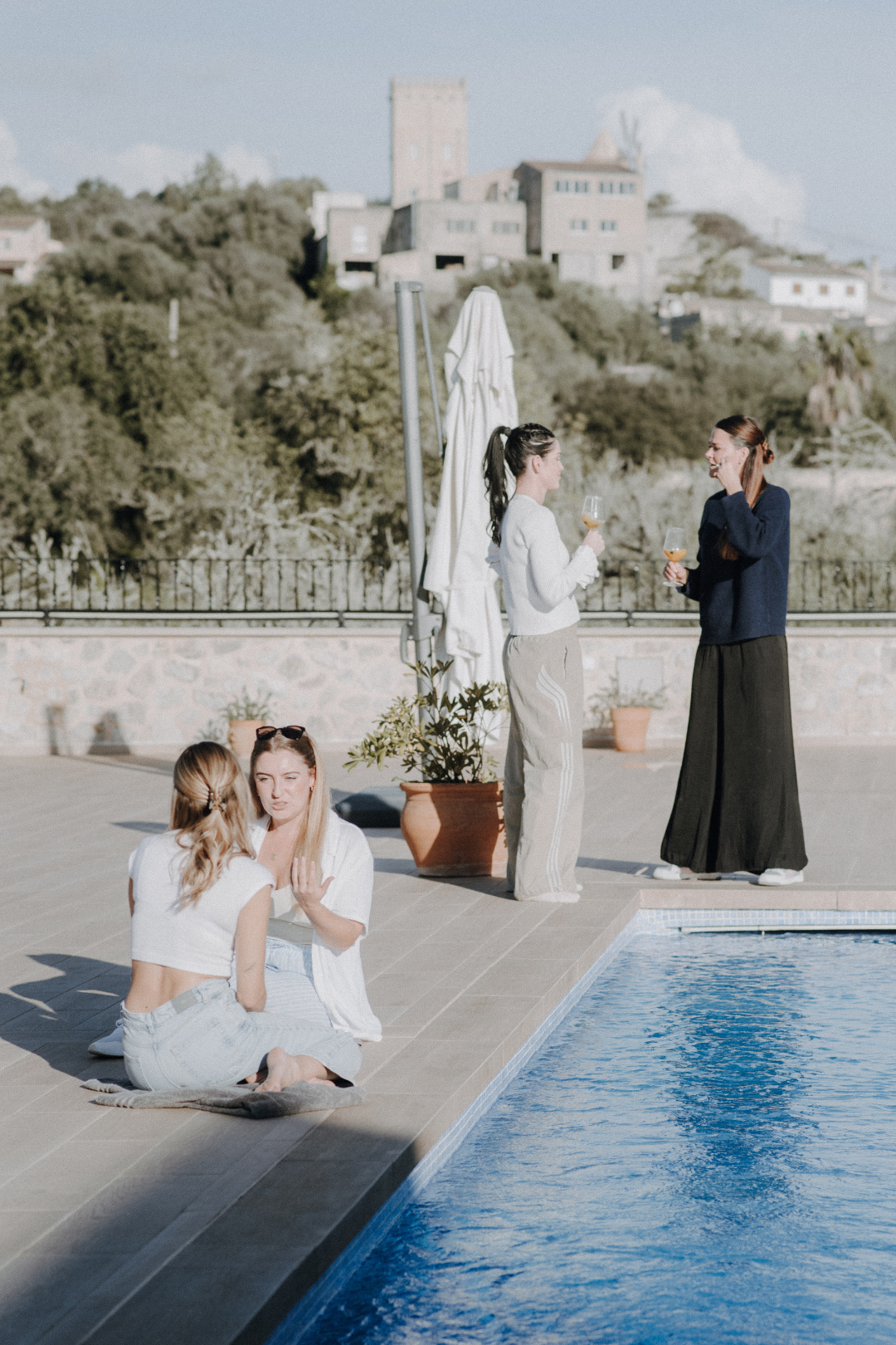 Vier Frauen sitzen und stehen am Pool, trinken Wein und unterhalten sich in einer sonnigen, ruhigen Umgebung mit einem Gebäude auf einem Hügel im Hintergrund.