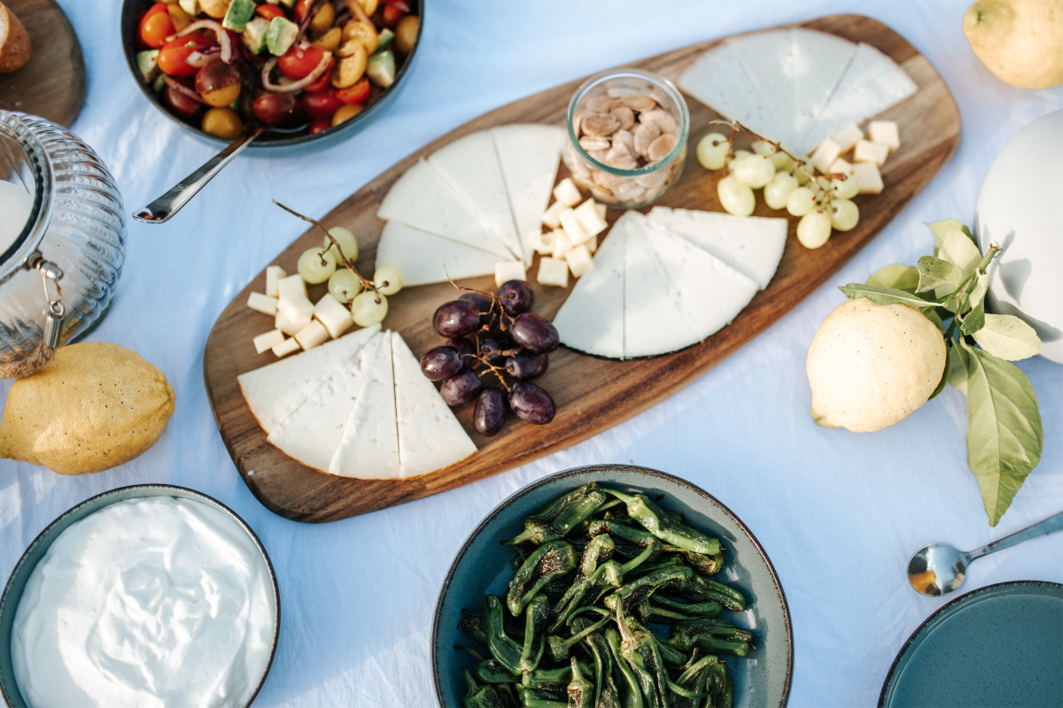 Eine Käse- und Obstplatte mit verschiedenen Käsesorten, Weintrauben, Nüssen, einem Zitronen, einer Schüssel mit grünen Peperoni, einer weiteren Schüssel mit Joghurt sowie einer Obstschale mit Cherry-Tomaten, roten Zwiebeln und Basilikum. Ein weißer T