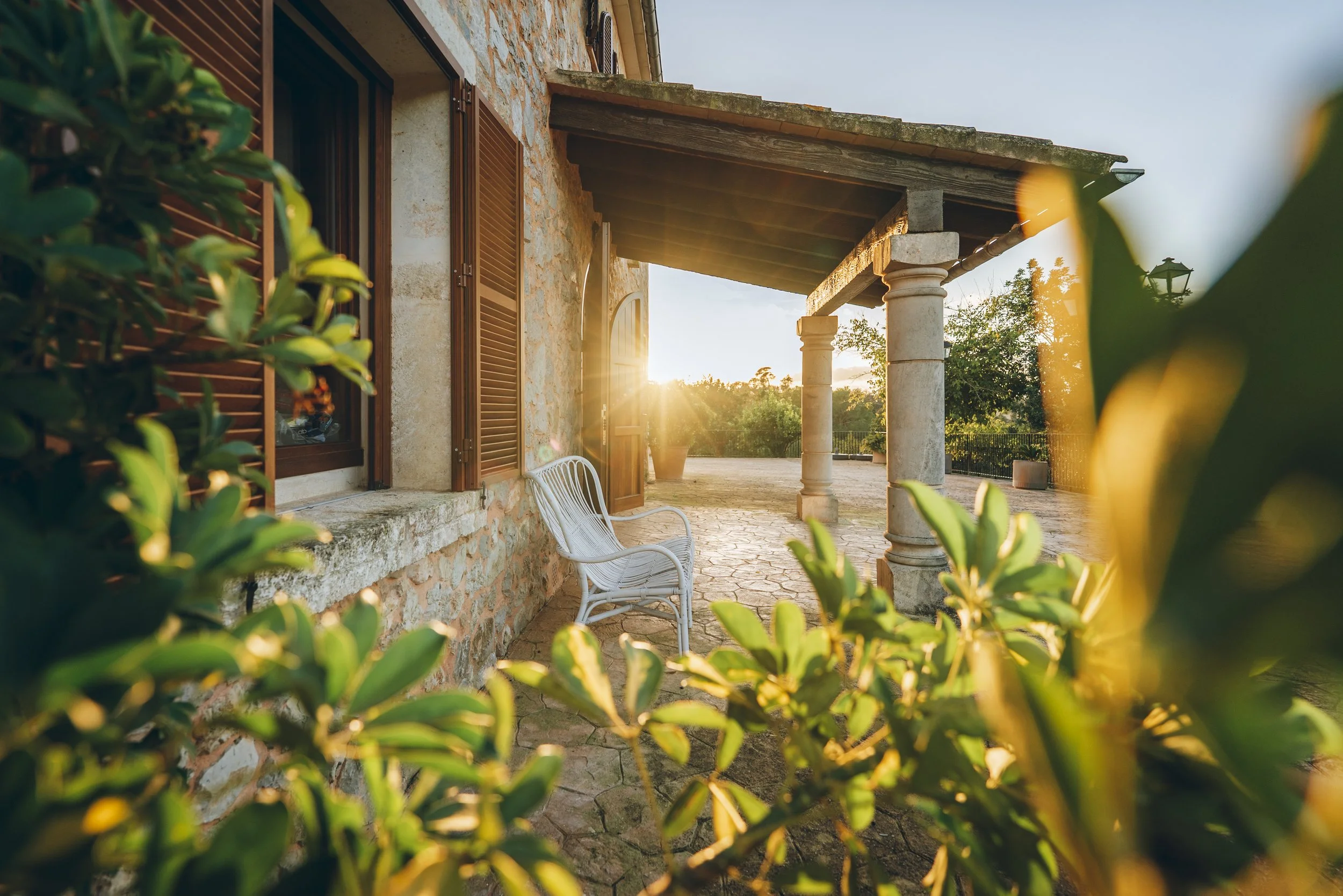 Sonnenuntergang über einer Veranda mit Steinsäulen, einer weißen Gartenbank, einer Steinmauer mit offenen Holzfenstern, umgeben von grünen Pflanzen.