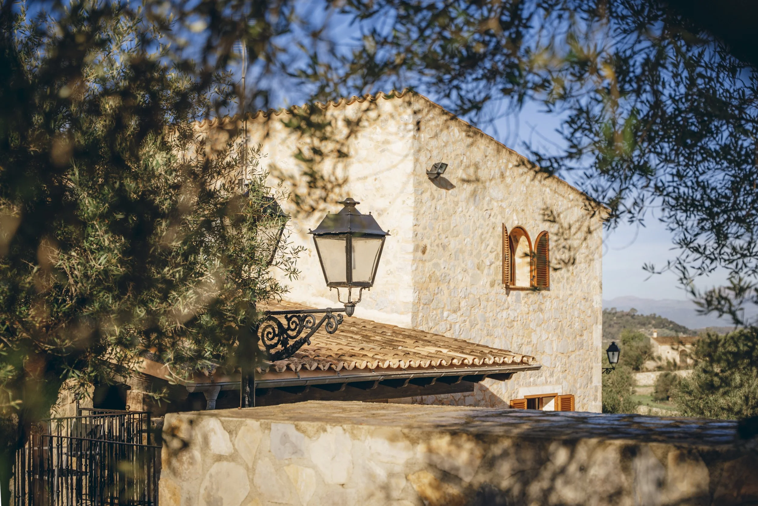 Ein Haus mit Steinfassade, braunen Fensterläden und einem Ziegeldach, umgeben von Bäumen, bei Sonnenuntergang.