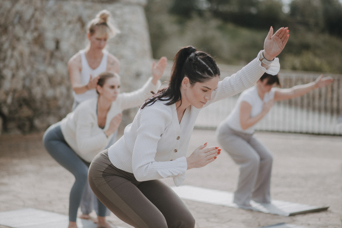 Vier Frauen machen Yoga im Freien auf Yogamatten, die Arme ausgestreckt und in einer tiefen Haltung.