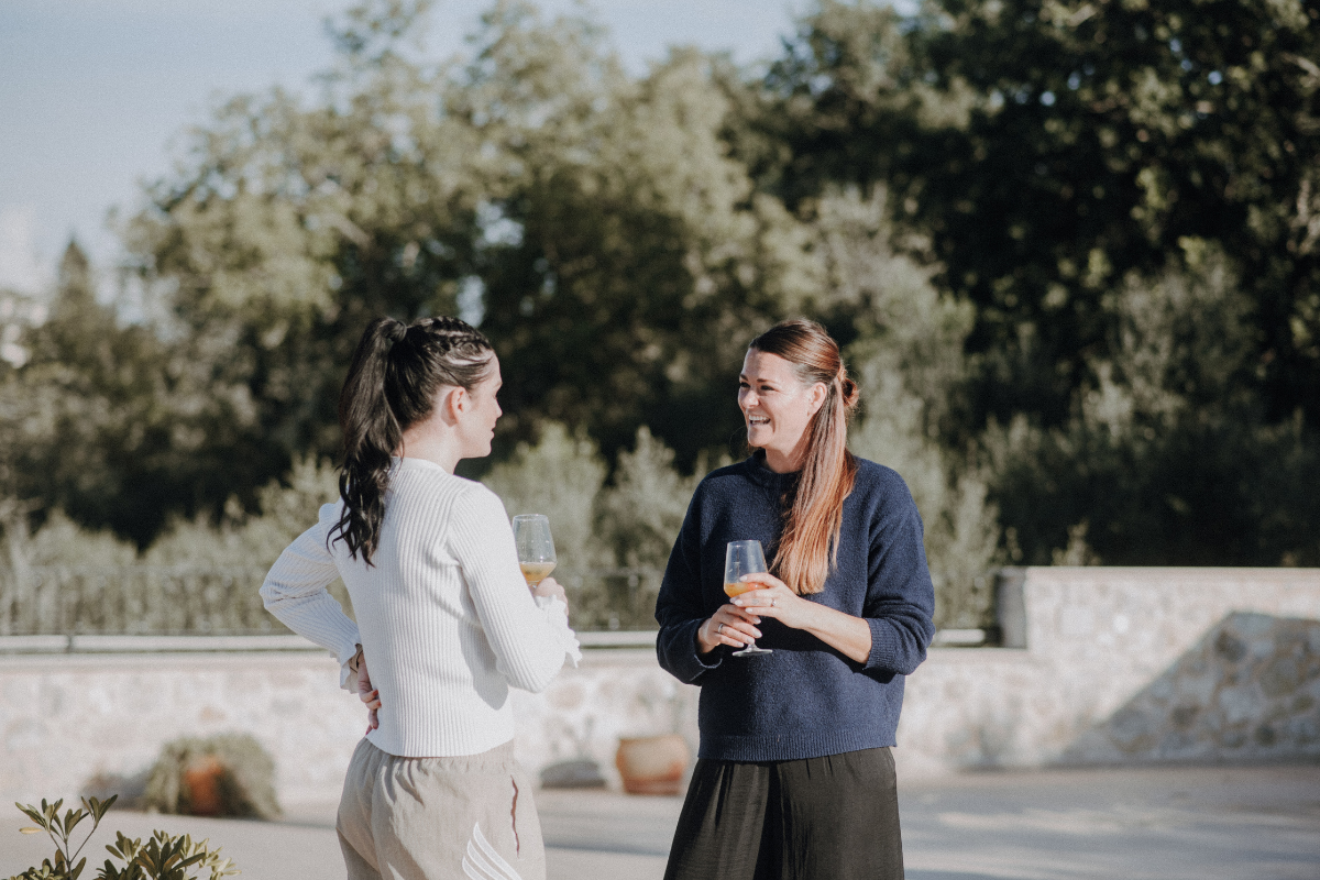 Zwei Frauen stehen im Freien bei einer Unterhaltung, jede hält ein Glas Wein, im Hintergrund Bäume und eine Steinmauer.