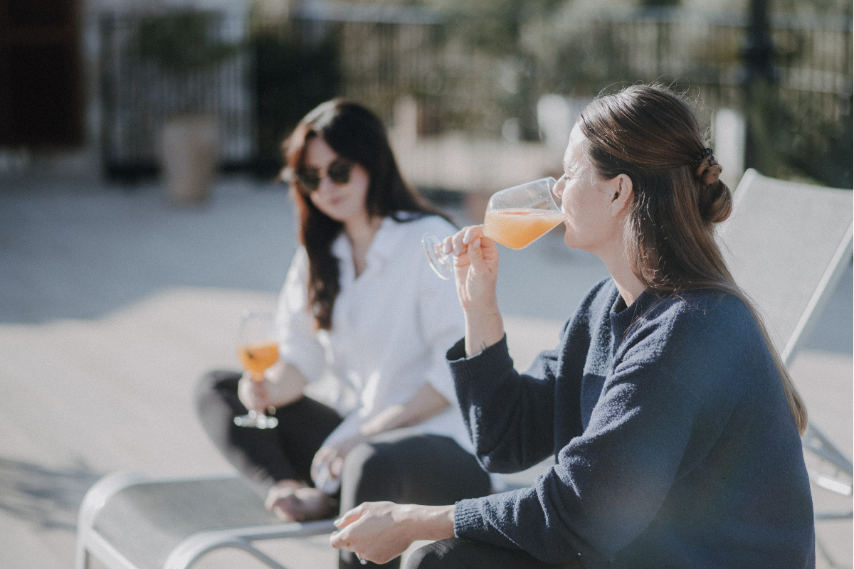 Zwei Frauen sitzen im Freien auf Liegestühlen und trinken Getränke aus Weingläsern, während sie die Sonne genießen.