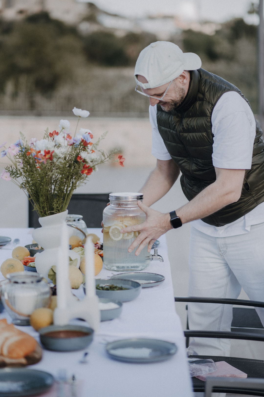 Ein Mann stellt Wasser mit Zitronenscheiben auf einem gedeckten Tisch mit Blumen und Essen auf einer Terrasse.