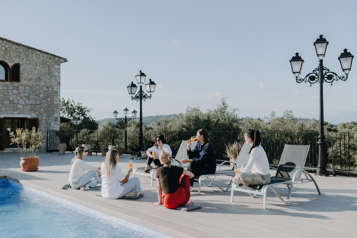 Eine Gruppe von jungen Leuten sitzt und entspannt an einem Pool auf einer Terrasse, trinkt und unterhält sich bei schönem Wetter.