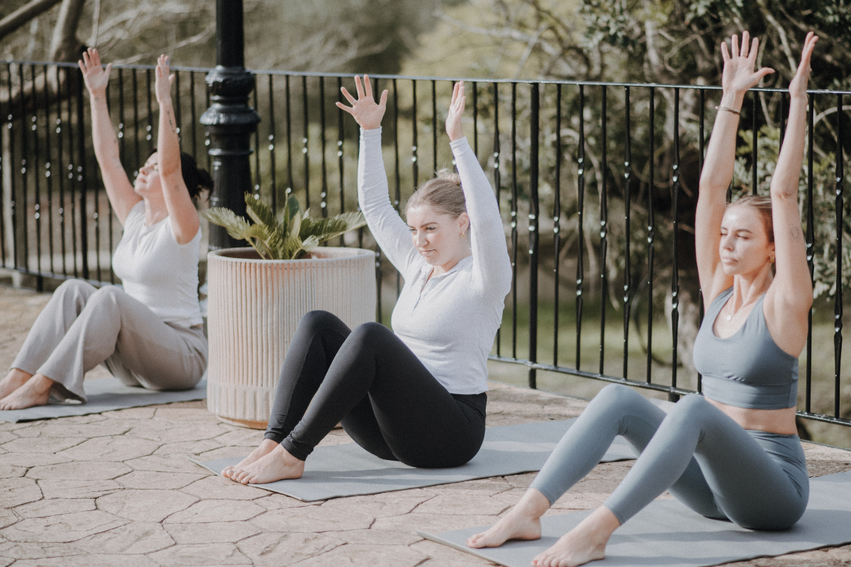 Drei Frauen machen Yoga im Freien auf Matten, sitzend mit erhobenen Armen, neben einem großen Pflanzkübel mit einer Pflanze, in einer parkähnlichen Umgebung.