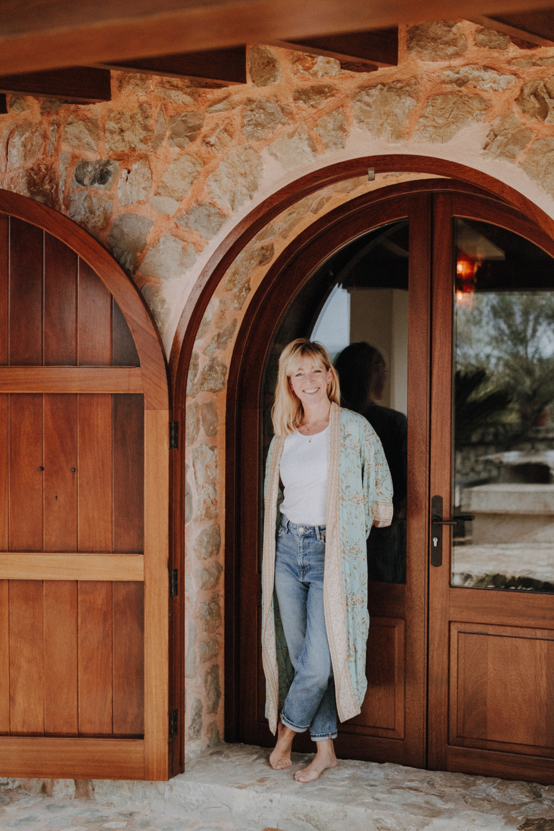 Frau steht barfuß am Eingang einer Stein- und Holztür, lächelt und trägt eine weiße Bluse, Jeans und einen langen, gemusterten Kimono.
