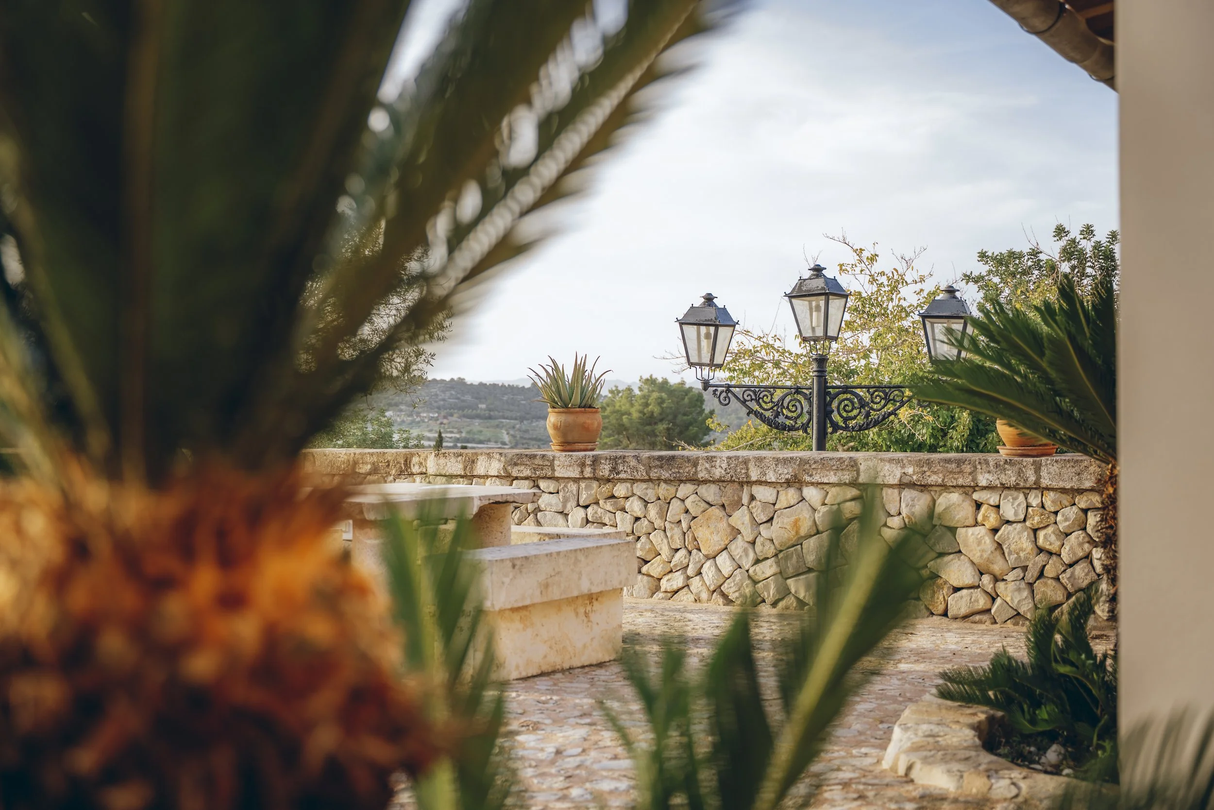 Blick auf einen terrassenartigen Außenbereich mit Steinmauer, Zierpflanzen in Tontöpfen, einen alten Laternenmast, umgeben von grünen Büschen und einem ziemlich weiten, hügeligen Landschaftshorizont
