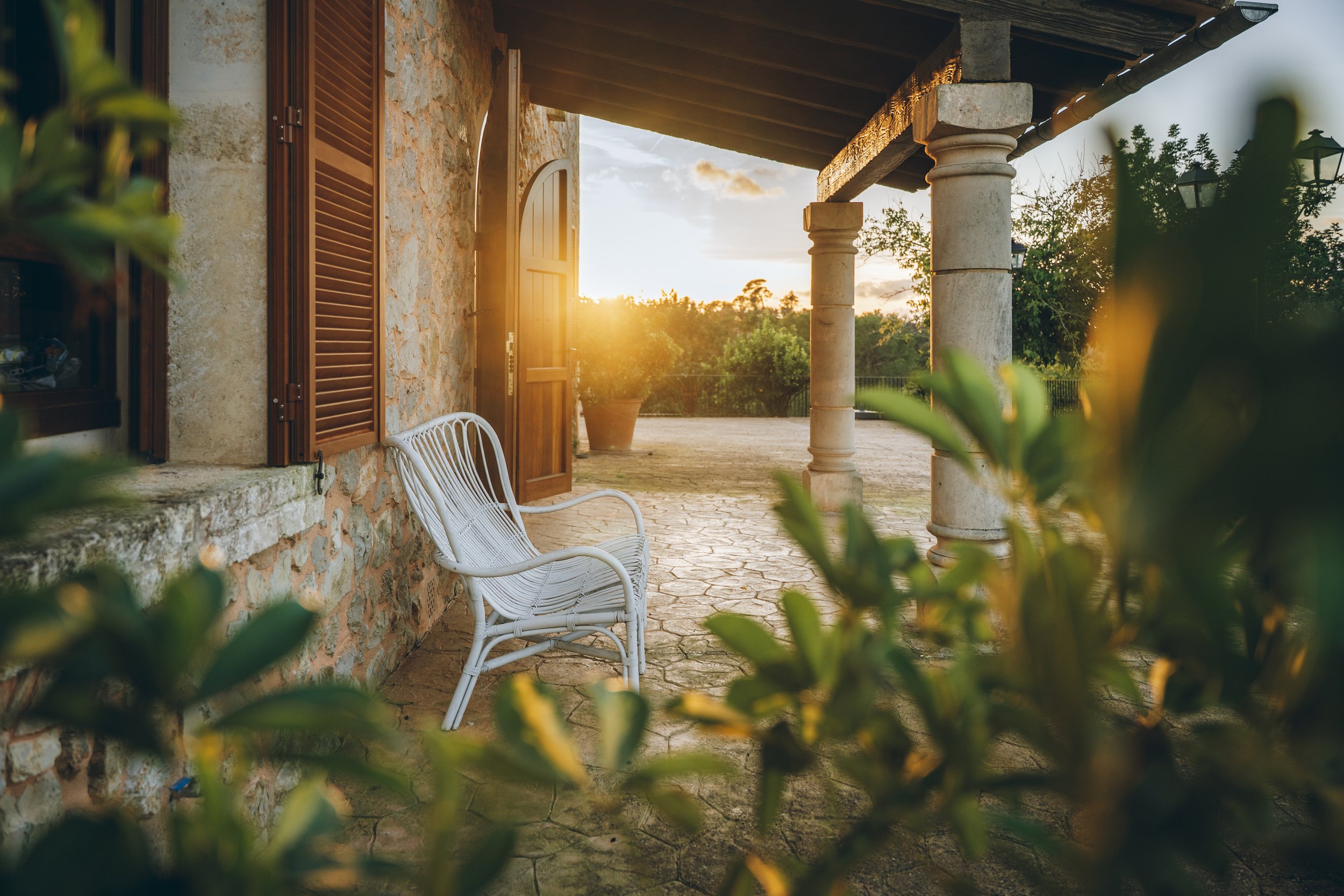 Ein ländlicher Hof im Sonnenuntergang mit einer weißen Gartenbank, einer Steinmauer und offenen Holztüren, umgeben von grünen Pflanzen und Bäumen.